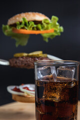 Delicious Burger with floating ingredients on the background of a wooden table. Bun with sesame seeds. Juicy meat cutlet. Carbonated brown drink with ice.