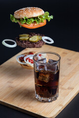 Classic Burger. Still life of a Burger and coke. Drink in a tall glass with ice. The components of the Burger are flying in the air on a black background. Meat cutlet, onion, tomato, lettuce and chees