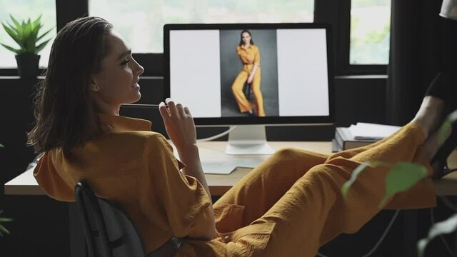 beautiful girl with makeup, dark hair and a yellow linen suit sits in front of a computer putting her feet in shoes on the table and looks at her photos from a photoshoot in the same outfit 