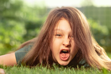 Crazy angry kid girl shouting with opened mouth and mad hair lying on the green grass on outdoor summer background. Closeup