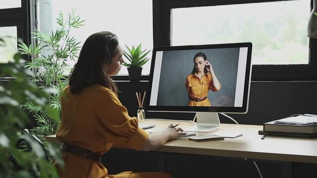 beautiful girl with makeup, dark hair and a yellow linen suit sits in front of the computer in the room and looks at her photos from the photoshoot in the same clothes 