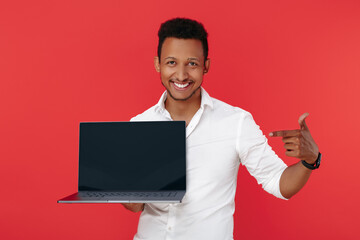 Happy african american young man holding laptop and pointing finger to screen over red background.