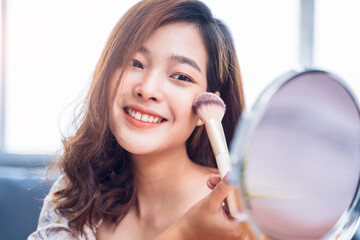 Asian beautiful smooth skin female professional beauty glamour cosmetics makeup tutorial using brush powder putting aesthetics on face looking into mirror living room modern white home lifestyle