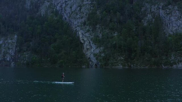 Aerial view moving forward side view, man wearing a cap paddle boarding on the lake, Scenic view green lake and green trees on the side of the mountains.