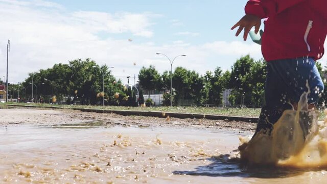 Kid jumping repeatedly in a muddy puddle puddly splashing water in slow motion. Carefree boy wearing red rain coat  and rubber boots having fun and enjoying rainy day. Childhood happy memories