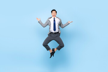 Full length playful portrait of happy ecstatic young Asian businessman jumping in mid-air doing wacky fun gesture in isolated studio blue background