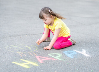 Fototapeta premium Little girl with syndrome down draws with chalk on the asphalt on summer day