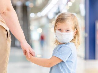 Little girl wearing medical protective mask holds mom's hand in a public crowded place - in a shopping mall or airport