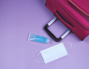 COVID-19 protection while traveling and new normal concept.Top view or flat lay of pink suitcase  with surgical face mask and alocohol sanitizer gel  on purple background