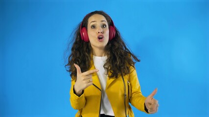 Naklejka premium Young woman in yellow jacket dancing while listening music on headphones in Studio with blue Background.