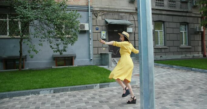 Happy woman in yellow dress and hat dancing alone on an empty city street listening to music on smartphone.