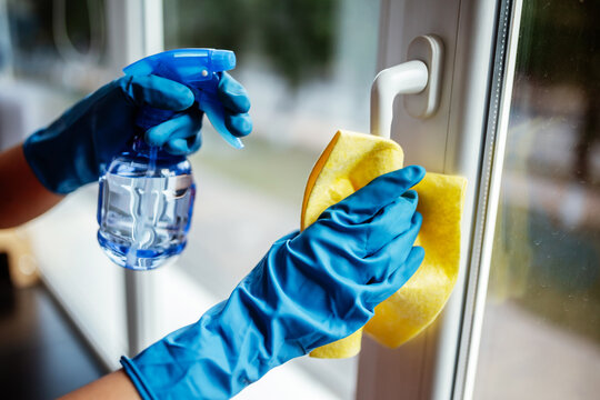 Closeup of woman wearing blue rubber gloves cleaning the window handle with microfiber rag and sanitizing spray during coronavirus pandemic quarantine. Health care and adaptation concept.