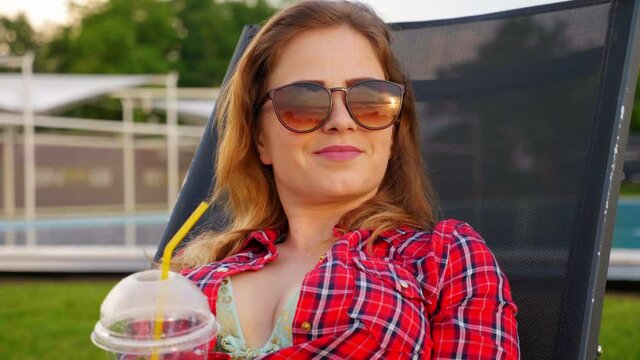 Woman in sunglasses lies on a deck chair against the backdrop of the pool, looks at the sunset, holds a plastic cup with a straw in hand, shakes head, smiles, rests. 4K slow motion footage