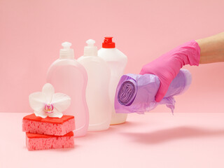 Bottles of dishwashing liquid, sponges and hand with garbage bags on a pink background.