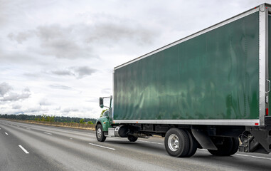 Big rig green semi truck transporting cargo in long box trailer running uphill on the wide highway...