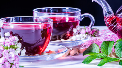Fototapeta na wymiar A mug of red tea in white hibiscus flowers and green leaves of medicinal tea on a wooden stand.Zen tea ceremony. Photo of red herbal Indian healing tea. Elegant mugs with a relaxing and tonic drink