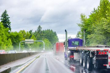 Blue big rig day cab semi truck with flat bed semi trailer driving on the wet dangerous road with rain dust running in convoy with another semi truck