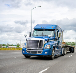Blue big rig semi truck with flat bed semi trailer running on wide road to warehouse for pick up load for next delivery