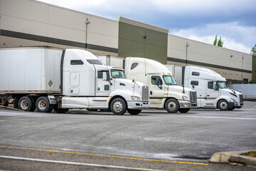 Different models of big rigs semi trucks with dry van semi trailers loading cargo at warehouse docks with rain weather