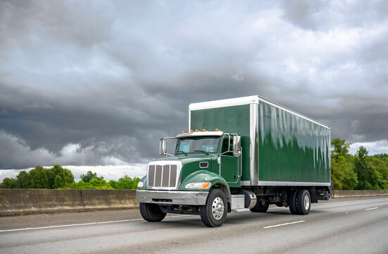 Small powerful green rig semi truck with box trailer running on the highway road with stormy sky on the background