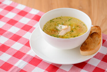 Chicken soup with vegetables and meat served in a white bowl over rustic wooden background with red plaid tablecloth.