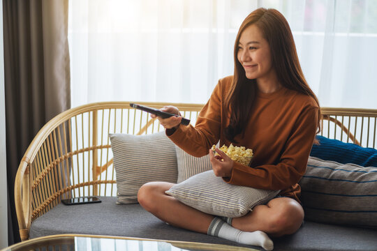 Close up image of a beautiful young woman eating pop corn and searching channel with remote control to watch tv while sitting on sofa at home