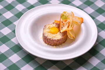 Close up of beef tartar with capers and small toasts. Served on a white plate over green plaid tablecloth background.