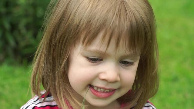 Close up of little girl in summer park sits on green lawn looks at camera, smiles and laughs. Outdoors. Happy child plays in nature