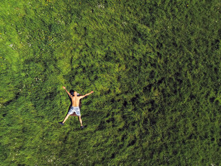 Bald man with naked torso sunbathing on a green grass in a field, legs and arm apart , Summer time. Aerial drone view.