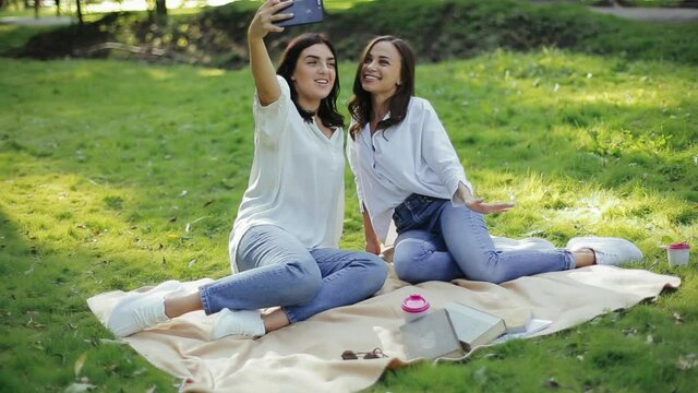 Pair of pretty european girls sit on grass on blanket in park, take bright selfie making funny grimaces. Two beautiful funny girl actively enjoy outdoor activities in their free leisure time in nature