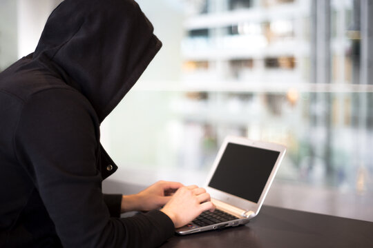 Anonymous hacker in black hood with laptop in the bussiness center. Light background. Cyber security.