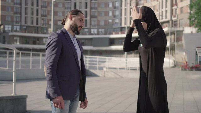 Angry young muslim woman shouting at bearded confident Middle Eastern man. Side view of husband and wife arguing outdoors on sunny day. Jealousy concept, relationship problems, conflict.