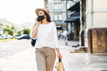 Obraz premium A young woman in a protective mask walks along a city street with a paper shopping bag in her hands and speaks on the phone. City walking and shopping after quarantine