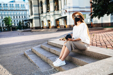 Fototapeta na wymiar Tourist girl with a protective mask on her face in casual clothes sitting on the steps in the city with a camera in her hands