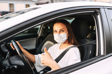 Protect yourself. Young woman with medical protective mask on her face is driving a car, she looks into camera and shows thumbs up. View from outside