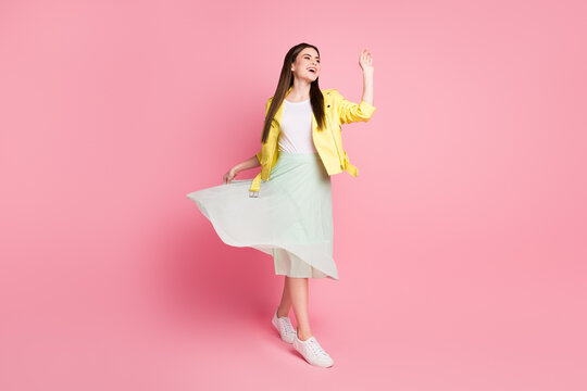 Full length photo of funky lady walk down street good mood raise green long wavy skirt wave arm see friends wear yellow leather jacket sneakers isolated pastel pink color background