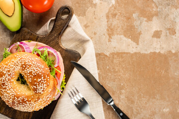 top view of fresh delicious bagel on wooden cutting board on aged beige surface with vegetables, cutlery and napkin