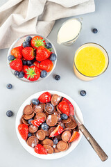 Trendy small chocolate cereal pancakes served with condensed milk, strawberry, blueberry and orange juice, in  white bowl, vertical, top view