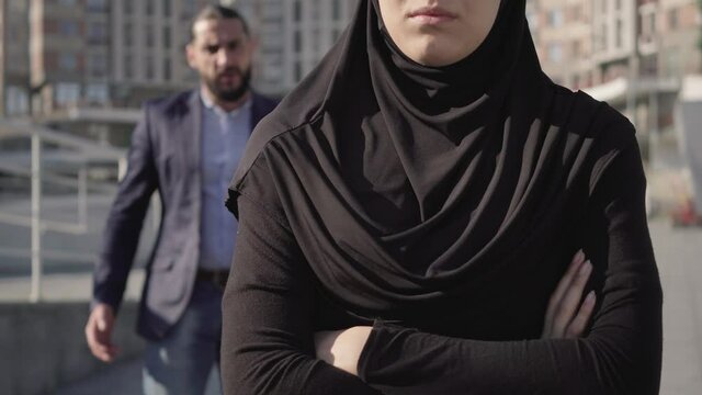 Unrecognizable muslim woman in traditional clothes standing with hands crossed as blurred angry man yelling at the background. Relationship problems in Middle Eastern family. Jealousy concept.