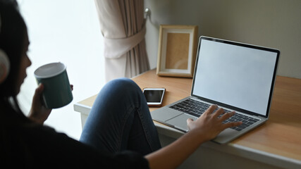 A woman is drinking a coffee while relaxing with a white blank screen computer laptop on the table.
