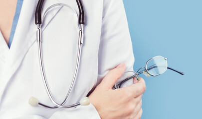 Hand of female doctor with stethoscope holds pair of glasses with thin metal frame. Minimal, blue background. Copy space. eye test checkups. Medicine mockup template