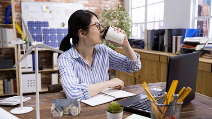 Casual working day of modern architect. Beautiful young Asian korean woman using laptop computer and drinking coffee cup while sitting at workplace. elegant lady engineer enjoy morning tea in office