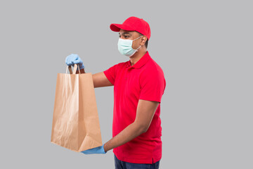 Delivery Man With Paper Bag in Hands Wearing Medical Mask and Gloves Watching Side Isolated. Red Uniform Indian Delivery Boy. Home Food Delivery. Paper Bag