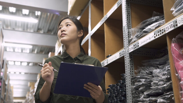 low to high portrait of warehouse worker woman looking up tall shelves doing inventory control in stockroom. female staff in uniform hold clipboard writing on paper while stock taking in storehouse