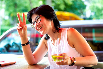A beautiful young brunette woman with glasses is sitting on a summer terrace with a glass of cocktail in her hand, openly smiling and showing her hands a gesture of peace, victory, having fun and enjo