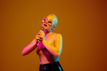 Musician. Young caucasian woman on brown studio background in neon light. Beautiful female model with microphone, speaker. Human emotions, facial expression, sales, ad concept. Freak's culture.