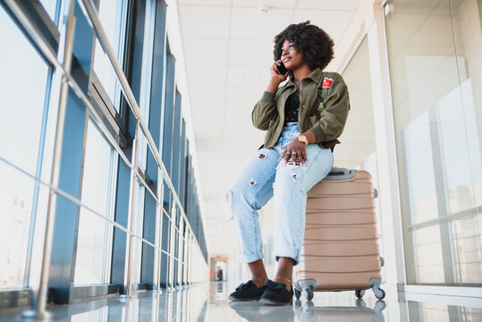 Portrait of happy young african american woman sitting on suitcase and talking with mobile phone at the station