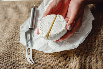 Cut Camembert cheese, French soft cheese with white mold in the hands of a woman on the background of a craft textile tablecloth, close-up. Knife for slicing cheese