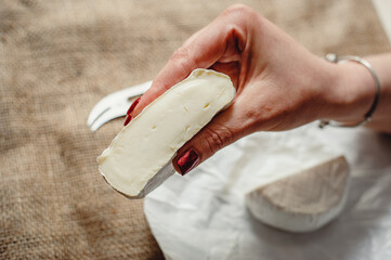 Cut Camembert cheese, French soft cheese with white mold in the hands of a woman on the background of a craft textile tablecloth, close-up. Knife for slicing cheese