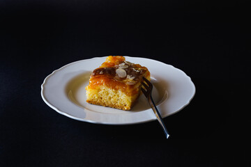 Slice of apricot cake on white plate with a dessert fork against black background
